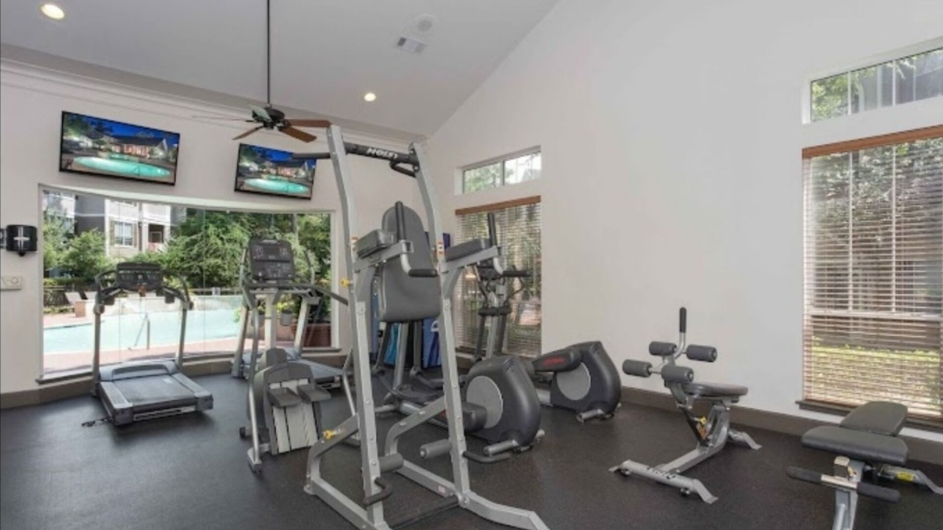 Ein Fitnessstudio mit unterschiedlichen Trainingsgeräten, großen Fenstern und einem Deckenventilator.