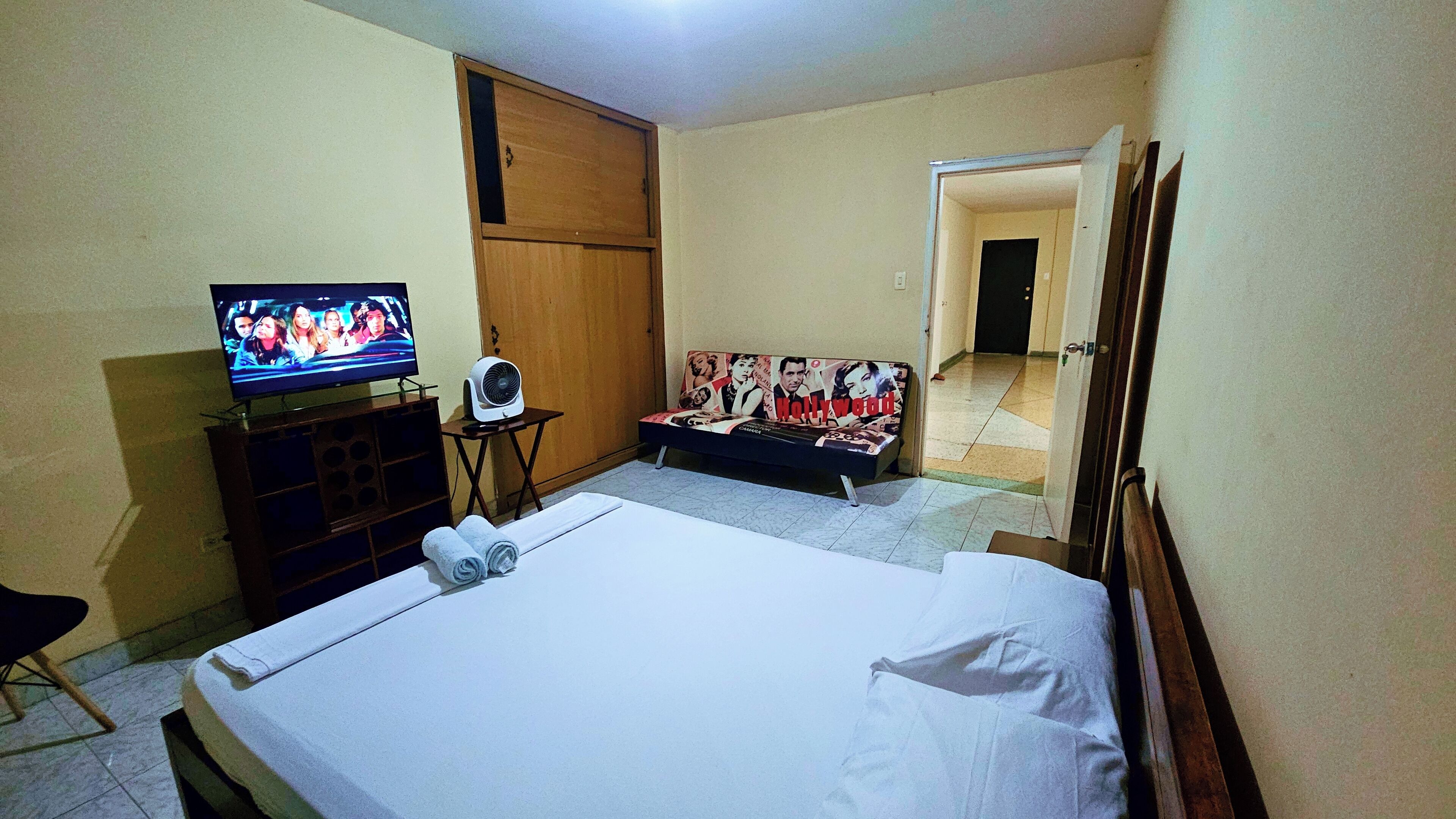 Een hotelkamer met een bed, een televisie, een klein tafeltje met een ventilator en een poster aan de muur.