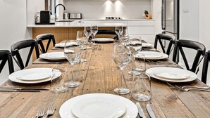 Ein Holztisch ist mit weißen Tellern, Weingläsern und Besteck in einer modernen Küche mit weißen Schränken und einer Edelstahl-Dunstabzugshaube gedeckt.
