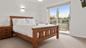 Ein Schlafzimmer mit einem hölzernen Bett, weißer Bettwäsche, einem Nachttisch, einer Lampe und Blick ins Freie durch eine Glastür.