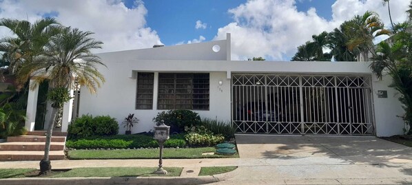 Un bâtiment moderne de couleur blanche, doté d’un grand portail de garage, et entouré d’un aménagement paysager soigné comprenant des palmiers et des arbustes.