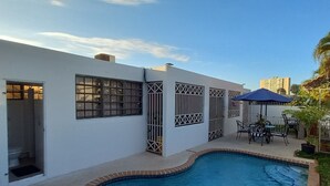 A swimming pool with a curved edge, a covered patio area with outdoor furniture, and a white building with barred windows. - San Juan, 2 rooms w/private pool, outdoor kitchen/BBQ & nearby & peaceful (San Juan)