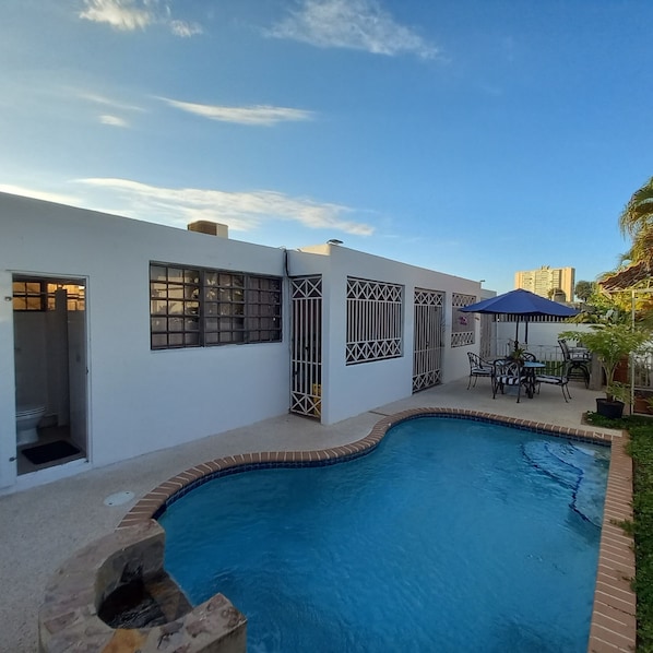 A swimming pool with a curved edge, a covered patio area with outdoor furniture, and a white building with barred windows.