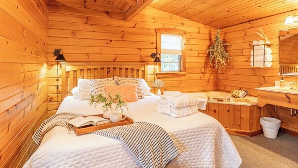 A wooden cabin bedroom with a bed, bedside tables, a window, and a bathroom with a sink and mirror.