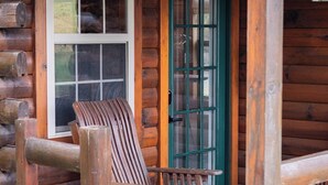 A log cabin with a wooden chair outside, a green door, and a window with blinds.
