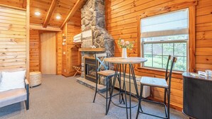 A log cabin interior with a stone fireplace, a round table, two chairs, a sofa, and a window with a view.