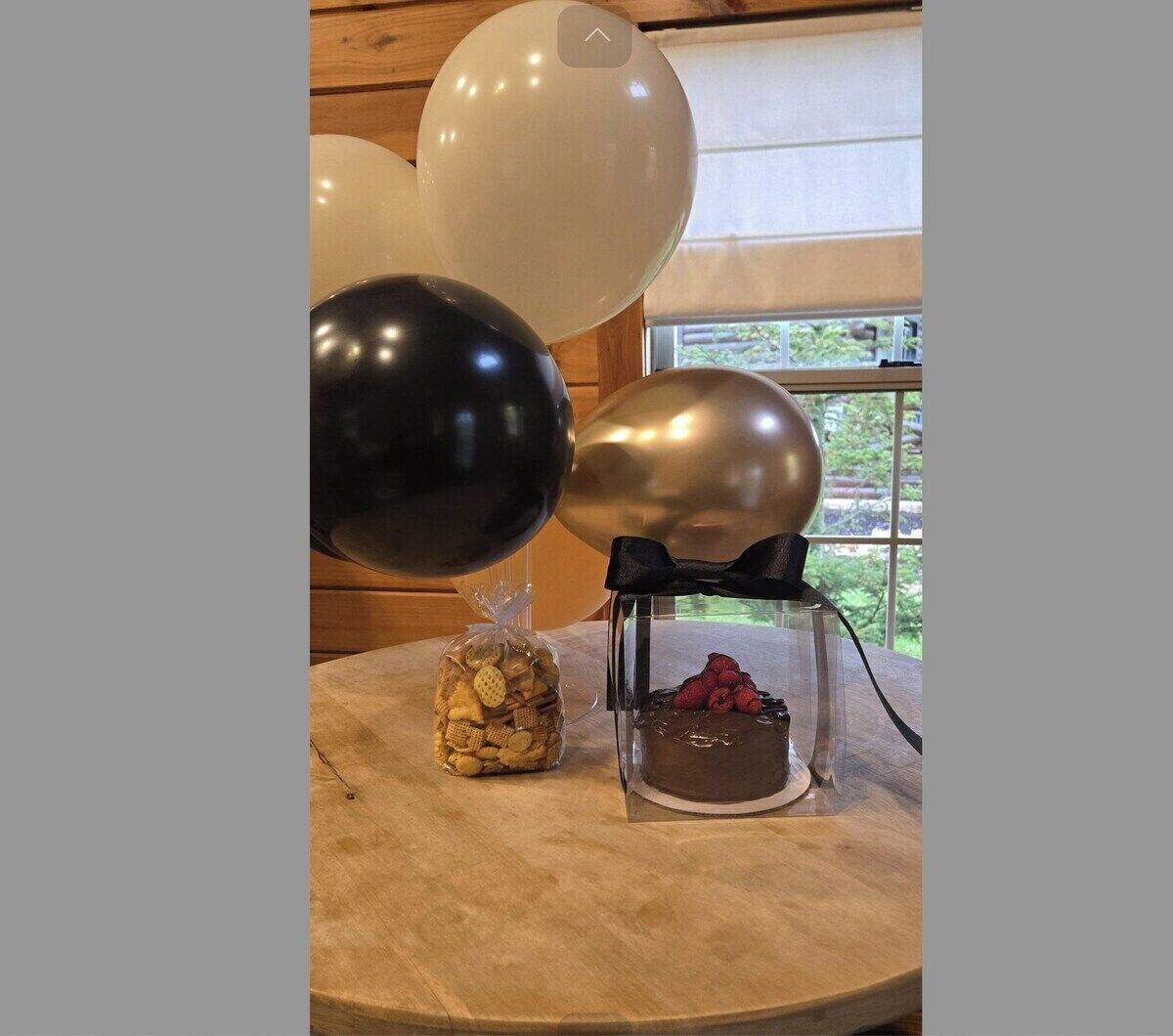 Una mesa con un recipiente de vidrio transparente que contiene un postre de chocolate cubierto de frambuesas, una bolsita de galletas y dos globos, uno negro y otro plateado, en una habitación con paredes de madera y una ventana.
