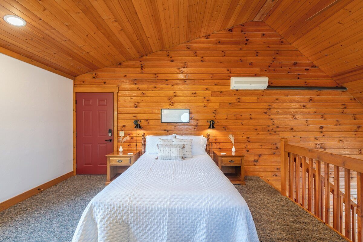 Une chambre avec un plafond en bois, un lit avec une literie blanche, deux tables de chevet avec des lampes, une porte rouge et un appareil de climatisation blanc.