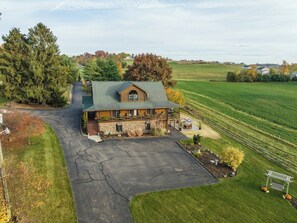 A large, two-story log cabin with a stone foundation, a covered porch, and a driveway leading to the entrance. - Spacious 6BR Retreat in Amish Country (Millersburg)