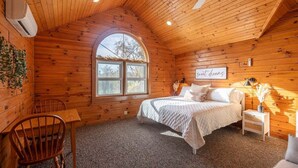 A cozy wooden cabin bedroom with a bed, a small table, a chair, and a window. - Spacious 6BR Retreat in Amish Country (Millersburg)