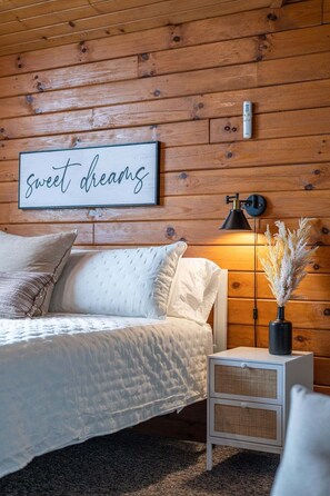 A bedroom with a wooden wall, a bed with white bedding, a bedside table with a lamp, and a sign that reads 'sweet dreams'. - Spacious 6BR Retreat in Amish Country (Millersburg)