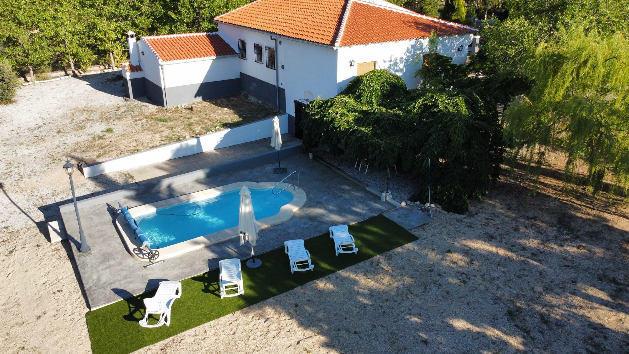 An aerial view of a house with a swimming pool, surrounded by greenery and a sandy area with lounge chairs.