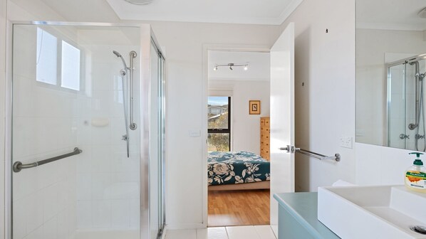 A modern bathroom with a glass shower enclosure, a white sink with a green soap dispenser, and a visible bedroom with a floral bedspread. - Cape Irvine - Linen not included (Cape Paterson)