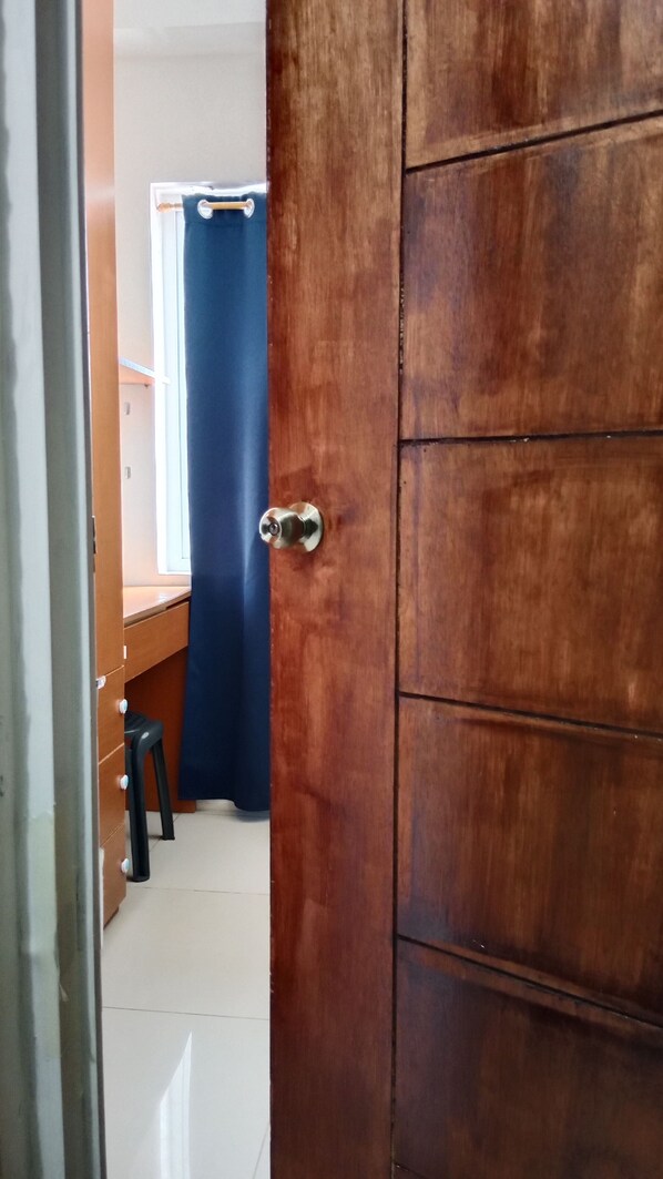 A wooden door with a metal handle, a blue curtain, and a small stool in a room with a tiled floor. - 1BR Haven in Royal Garden Residences (Cebu City)