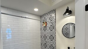 A bathroom with white tiled walls, a black and white patterned accent wall, a black shower area, a white sink with a black faucet, and a small shelf with toiletries.