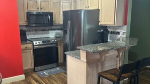 Una cocina compacta con refrigerador de acero inoxidable, microondas y estufa. Se observa una pequeña isla de madera con encimera de granito y un taburete de bar.