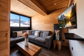 Ein gemütliches Wohnzimmer mit Holzbalkendecke, einem grauen Sofa mit dekorativen Kissen, einem Holztisch und einem großen Fenster mit Blick auf eine verschneite Landschaft.