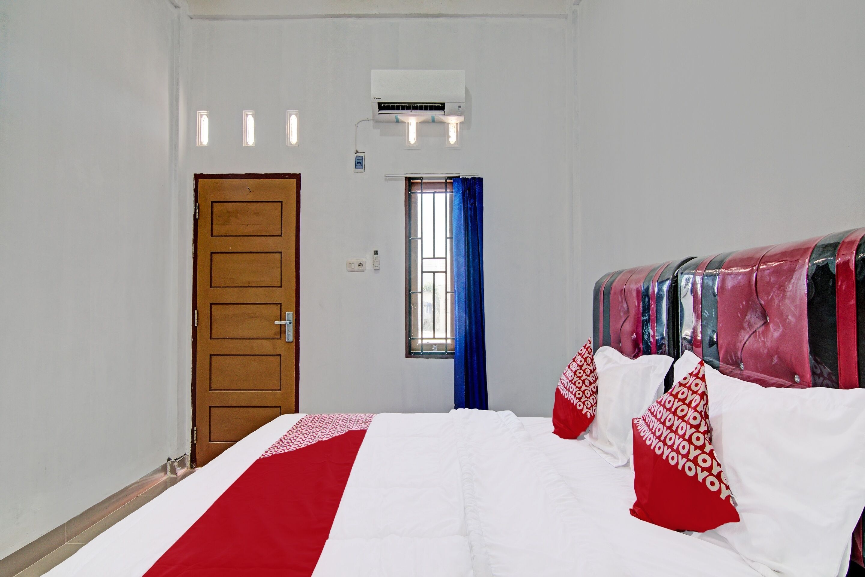 A hotel room with a bed, a window with blue curtains, an air conditioning unit, and a wooden door.
