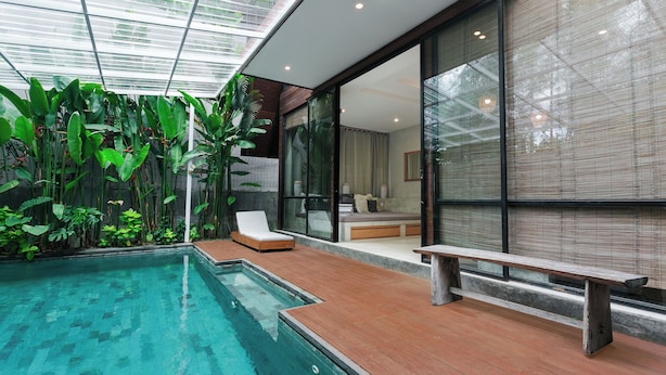 A modern indoor pool area with a wooden deck, a bench, and lush green plants. - Kalino Villa by Betterplace (Canggu)