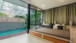 A modern bedroom with a large window, a bed with decorative pillows, a small table, and a view of a swimming pool. - Kalino Villa by Betterplace (Canggu)