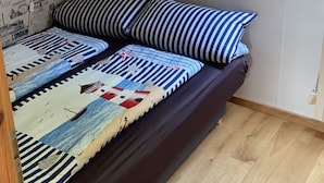 Ein gemütliches Zimmer mit einer dunkelbraunen Couch, gestreiften Kissen und einer farbenfrohen Decke. Eine kleine, grüne Pflanze hängt von der Decke, und das Zimmer hat ein geneigtes Dach mit einem Oberlicht.