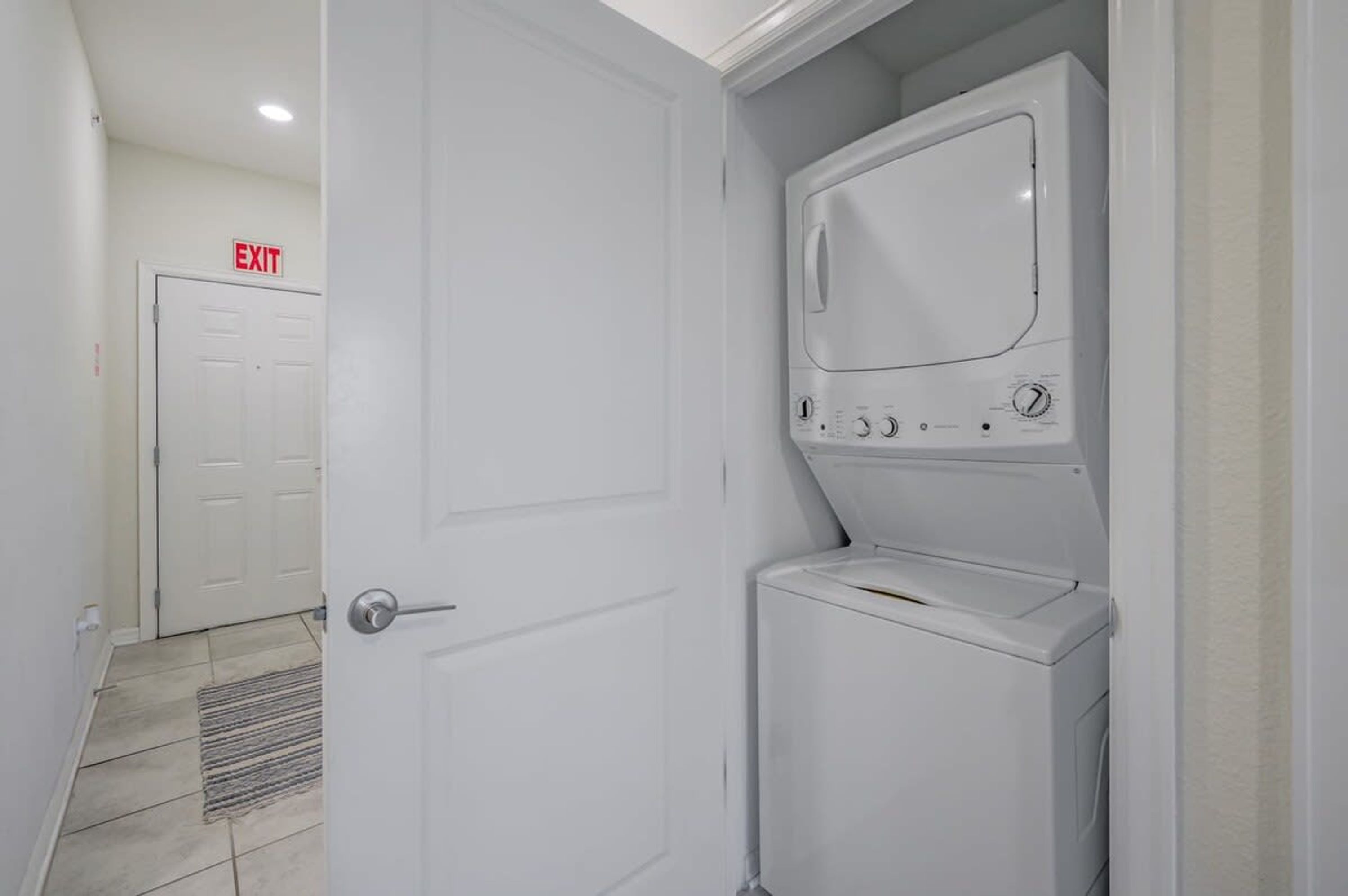 A laundry room with a front-loading washing machine and dryer, an exit sign, and a door leading to another room.