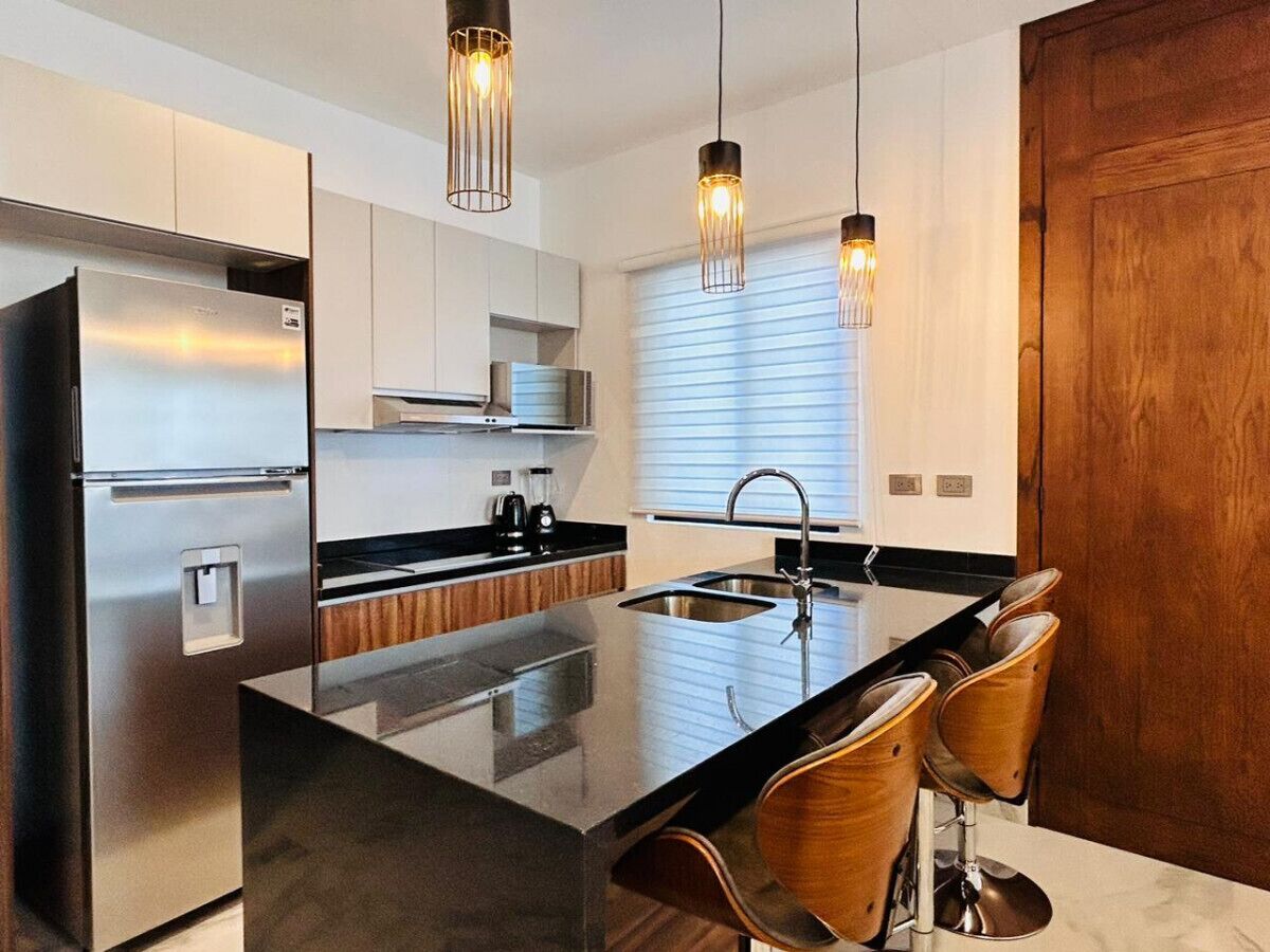 Una cocina moderna con refrigerador de acero inoxidable, una isla de encimera negra con taburetes de bar, iluminación colgante y armarios de madera.