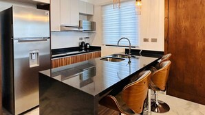 Eine moderne Küche mit Edelstahlkühlschrank, einer Kücheninsel mit schwarzer Arbeitsplatte und Barhocker, Pendelleuchten und Holdekor-Schränken.