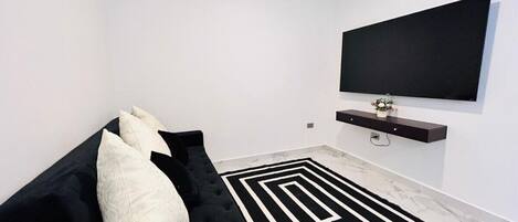 A modern living room with a black sofa, a black and white rug, a flat-screen TV mounted on the wall, and a small shelf with a flower vase.