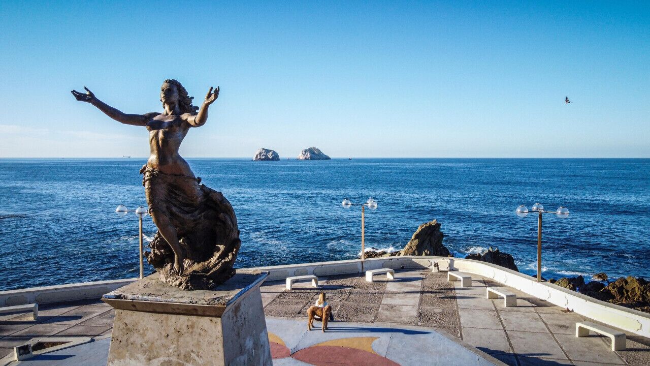 Eine Statue einer Frau mit ausgestreckten Armen, die auf das Meer blickt, mit Inseln in der Ferne und einem wolkenlosen Himmel.