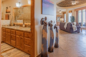 Un baño con un espejo grande, armarios de madera y un lavamanos. Se observan elementos decorativos en la pared y un ventilador de techo.
