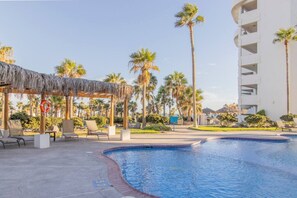 A pool area with lounge chairs, a thatched gazebo, palm trees, and a white building.