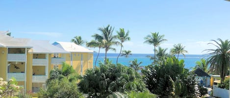 Un edificio amarillo con balcones, rodeado de vegetación tropical y vista al océano.