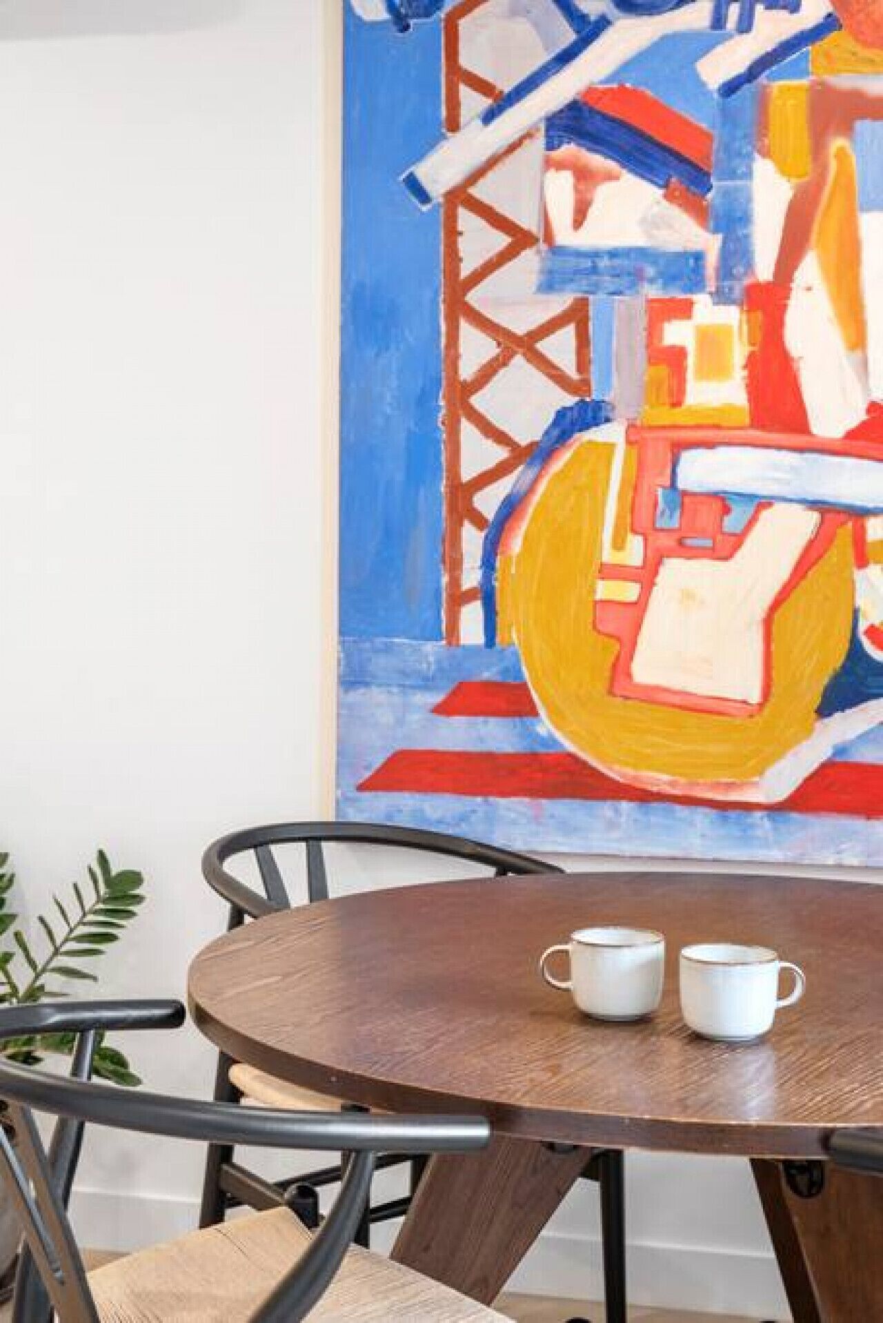 A round wooden table with two white cups, a metal chair, and a large abstract painting on the wall.