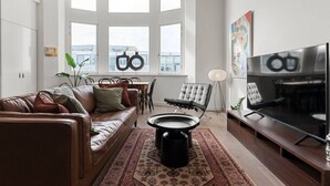 Un salon moderne avec un canapé en cuir marron, une table basse noire, un grand téléviseur à écran plat et un coin repas comprenant une table et des chaises blanches.