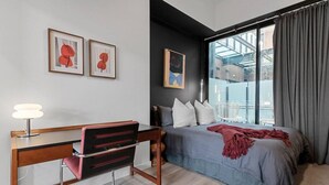 Une chambre moderne avec un lit, un bureau et une chaise. Les murs sont ornés d’œuvres d’art encadrées, il y a une fenêtre avec des rideaux et une vue sur l’extérieur.