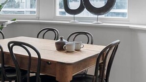 Un coin repas avec une table en bois et des chaises noires, une fenêtre donnant sur des immeubles et un miroir décoratif accroché au mur.
