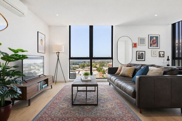 A modern living room with a large window, a leather sofa, a coffee table, and a TV.