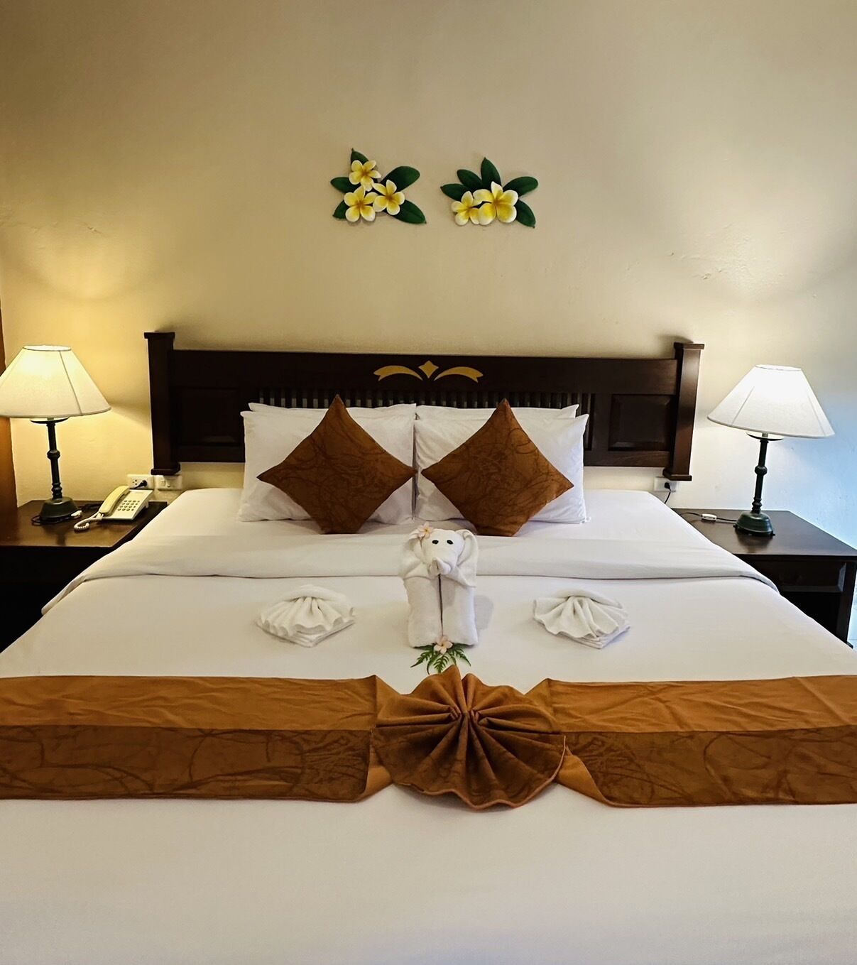 A neatly made bed with white linens, two brown decorative pillows, a small white towel arrangement, and a brown decorative centerpiece.