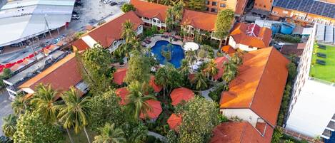 An aerial view of a resort with a central pool, surrounded by palm trees and buildings with red roofs.