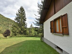 Uma casa com fachada branca e venezianas de madeira, rodeada por um gramado e árvores altas.