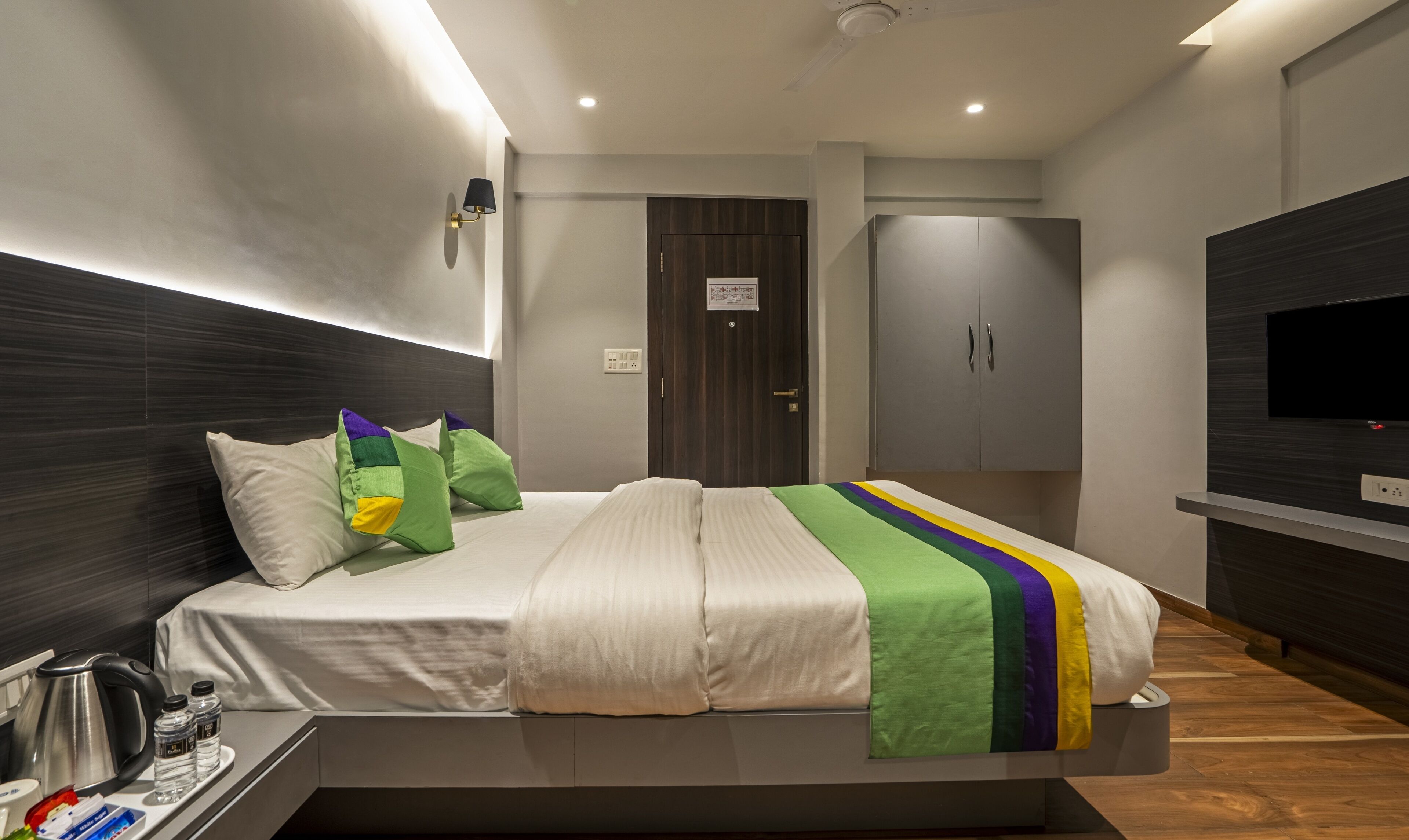 A modern hotel room with a large bed, a TV mounted on the wall, a wardrobe, and a bedside table with a kettle and water bottles.