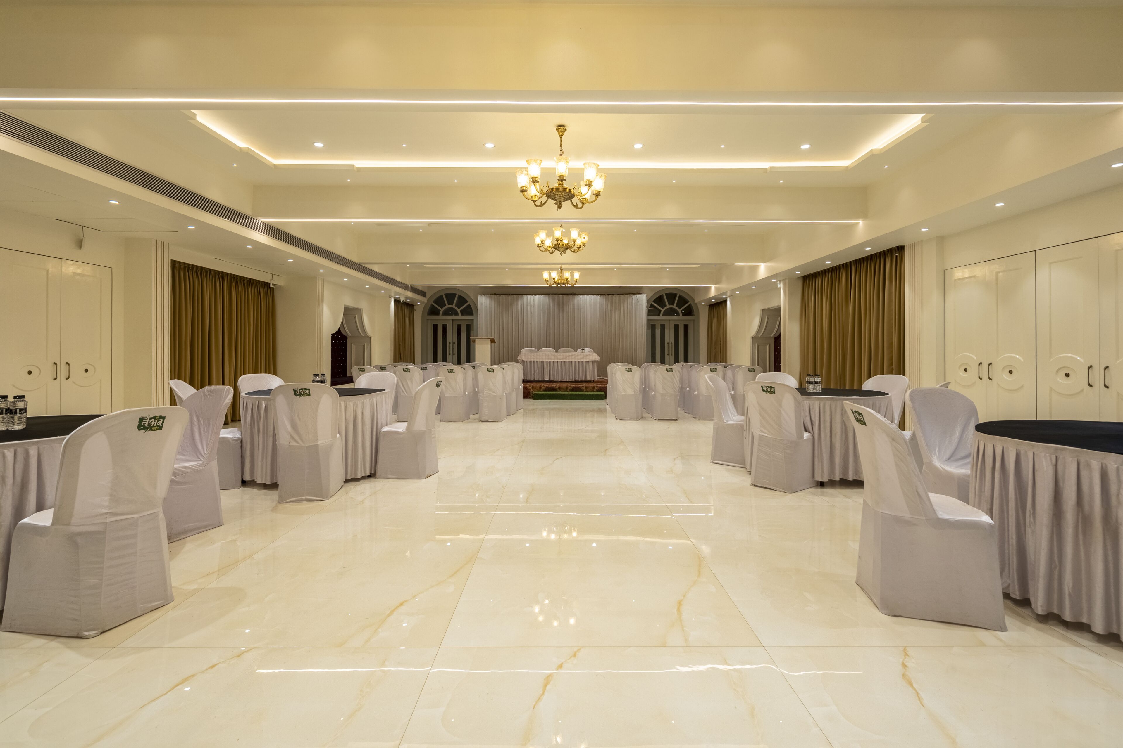 A spacious banquet hall with a polished marble floor, rows of white chairs with grey sashes, and a central stage with a backdrop.