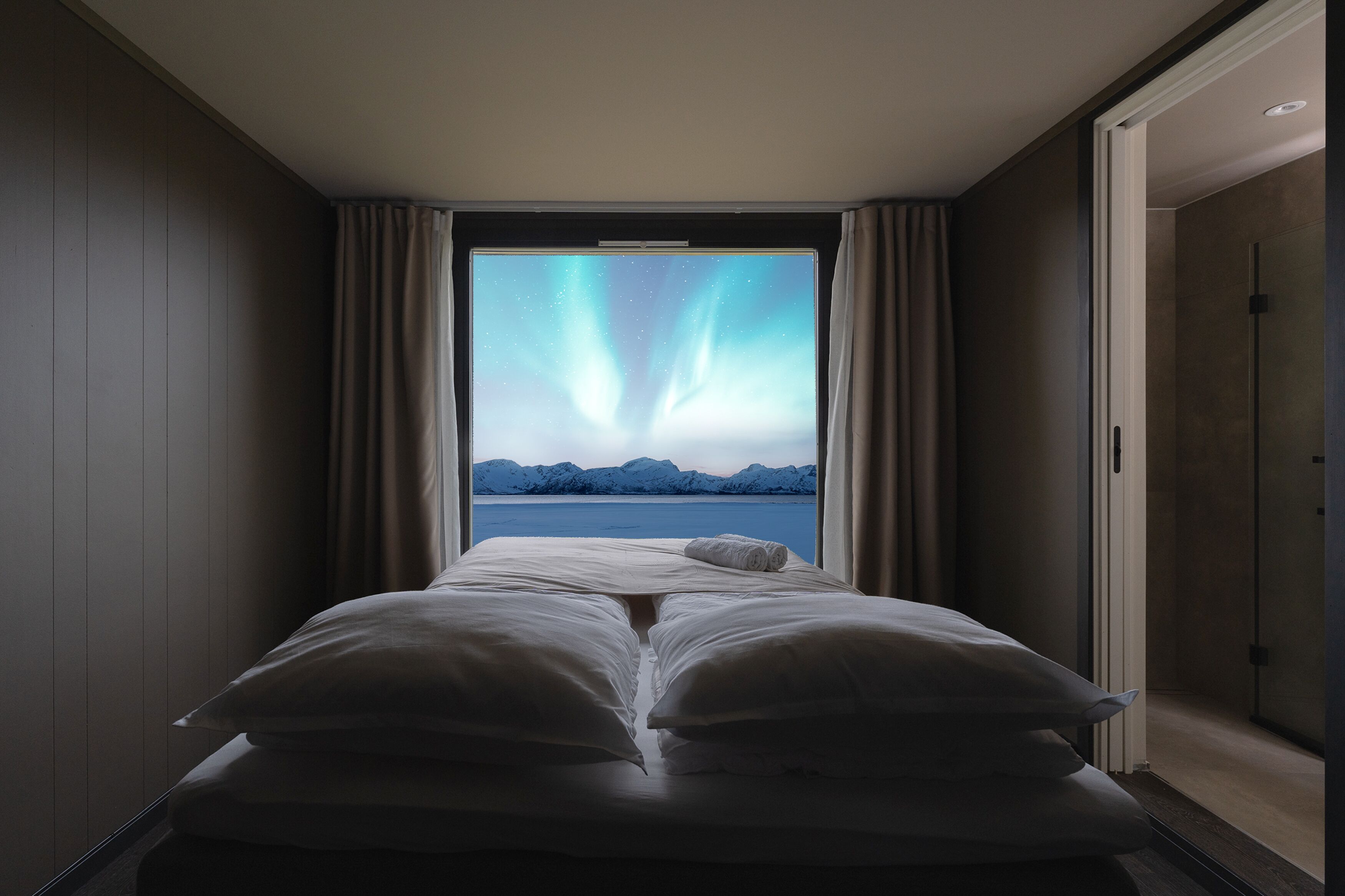 A bed with a view of the aurora borealis through a window, a door to the right, and a view of a body of water and mountains in the distance.