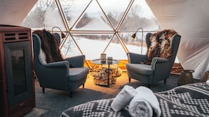 A cozy interior with two grey armchairs, a small round table, and a bed with a patterned blanket. - AERA - Aurora Glass Cabins (Tromsø)