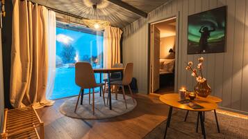 A cozy room with a dining table, chairs, a small round table with a candle and flowers, a large window with curtains, and a painting of the aurora borealis.