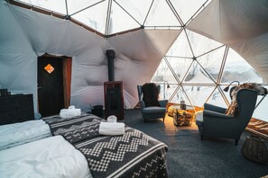 A cozy interior of a geodesic dome with a bed, seating area, and a fireplace. - AERA - Aurora Glass Cabins (Tromsø)