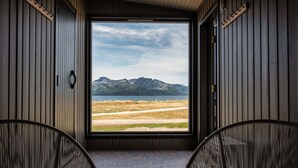 A room with a large window offering a view of mountains and a beach. There are two chairs in the room. - AERA - Aurora Glass Cabins (Tromsø)