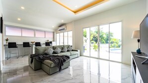 A spacious living area with a sofa, chairs, a TV, and a large window providing a view of the outdoors. - 4 Bedroom Private Pool Villa - OV10 (Hua Hin)