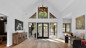 A spacious room with wooden flooring, a large chandelier, and a view through large glass doors to an outdoor area. - Luxury Lakeside Retreat w/ Panoramic Flathead View (Lakeside)